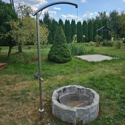 JS Welding & Renovation Jakub Siemieniec - Metalowy grill wiszący nad kamiennym paleniskiem, z mechanizmem regulacji wysokości na tle zadbanego ogrodu i błękitnego nieba z chmurami.