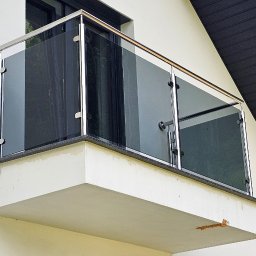 JS Welding & Renovation Jakub Siemieniec - Nowoczesna balustrada ze szkła i stali nierdzewnej na balkonie z kamiennym wykończeniem, widoczna z dołu pod kątem. Tło stanowi elewacja budynku i fragment dachu.