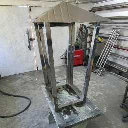JS Welding & Renovation Jakub Siemieniec - Metalowa konstrukcja przypominająca lampion, wykonana ze stali nierdzewnej, w warsztacie ślusarskim, w trakcie tworzenia. Widoczny spawacz i inne elementy warsztatu.
