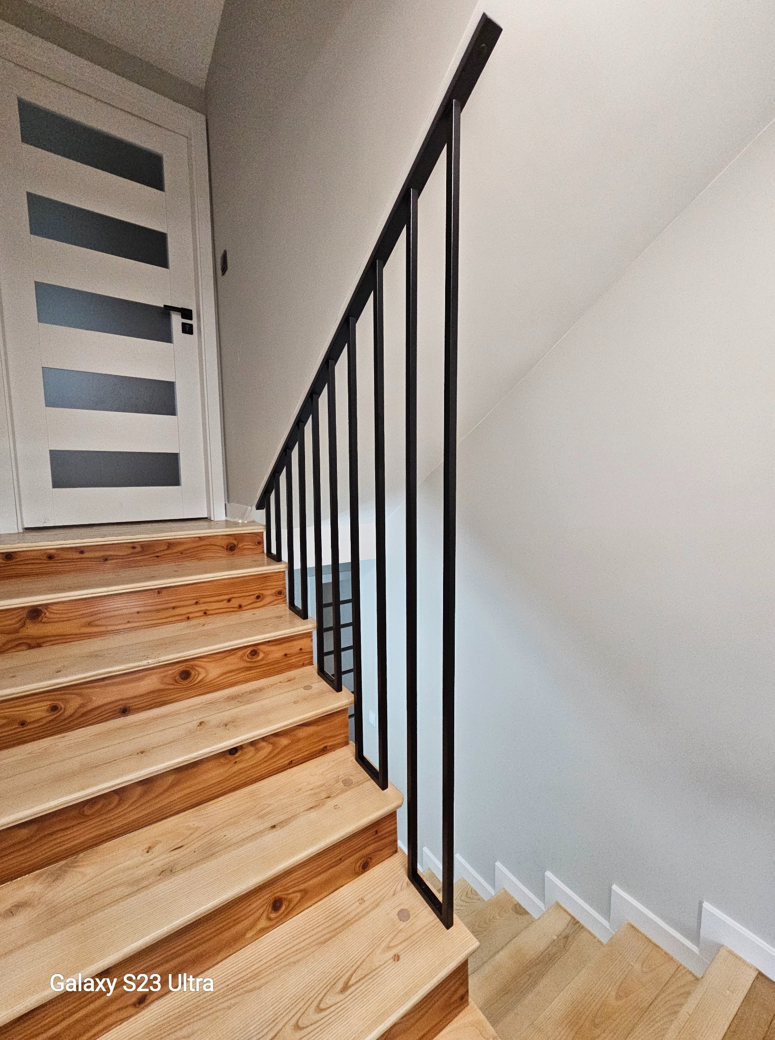 Nowoczesna, czarna poręcz metalowa na drewnianych schodach wewnątrz domu. Minimalistyczny design, białe ściany i drzwi z przeszkleniami. Ujęcie z dołu.