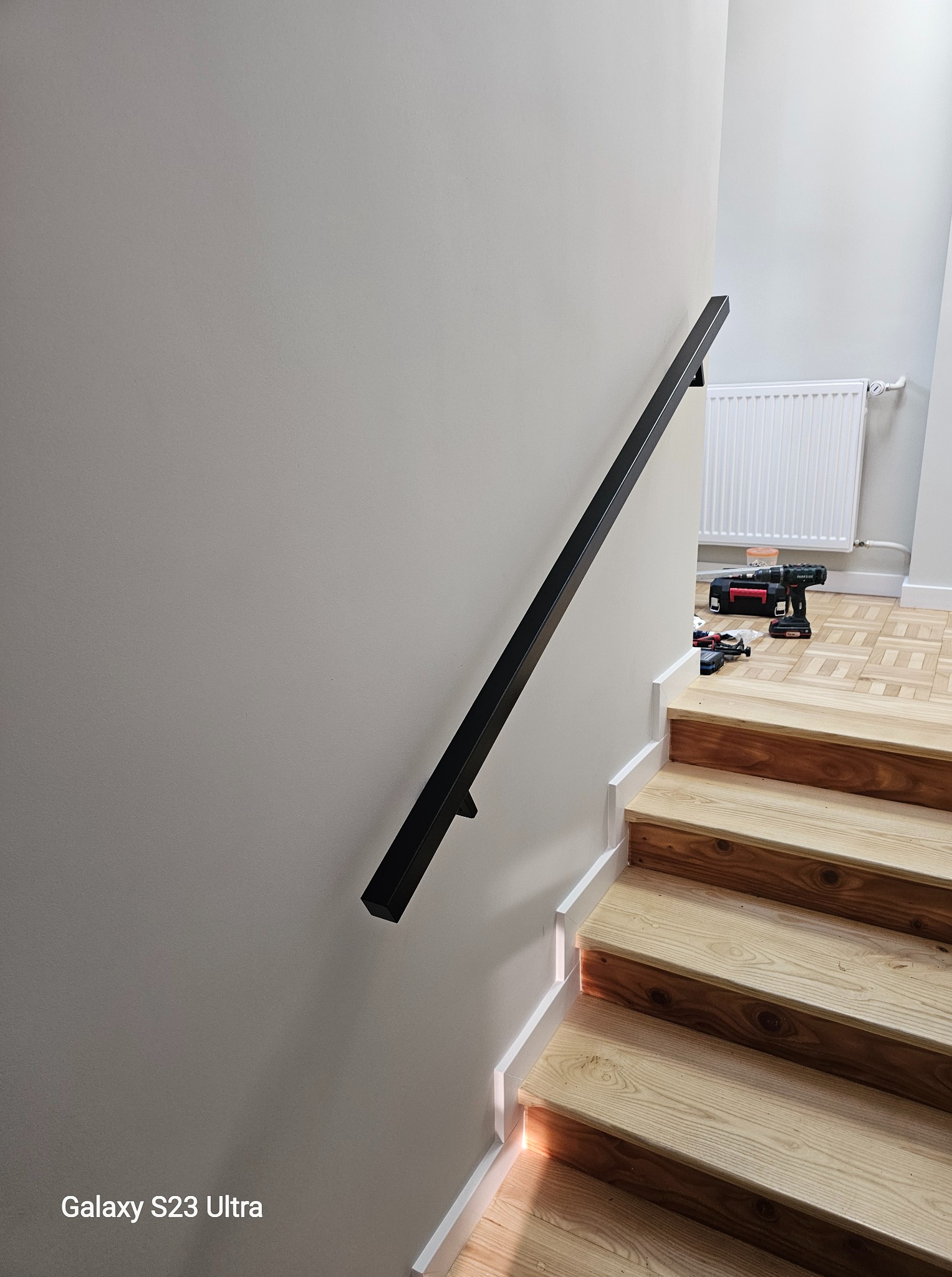 Czarna, minimalistyczna poręcz zamontowana na ścianie przy drewnianych schodach. W tle widoczne narzędzia i grzejnik. Ujęcie z dołu, podkreślające linię poręczy.