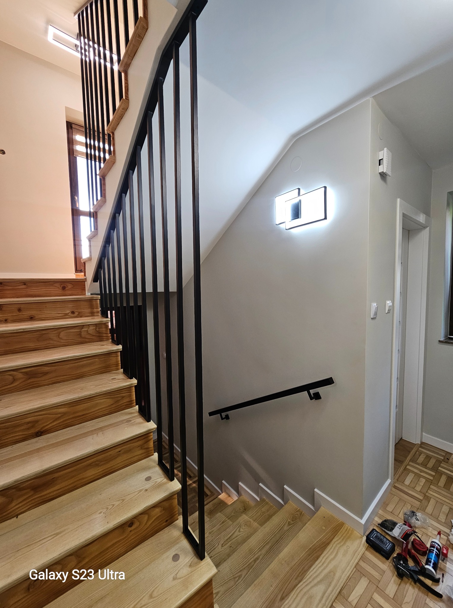 Drewniane schody z czarną, metalową balustradą o minimalistycznym designie. Widoczne narzędzia na podłodze sugerują trwający montaż lub prace wykończeniowe.
