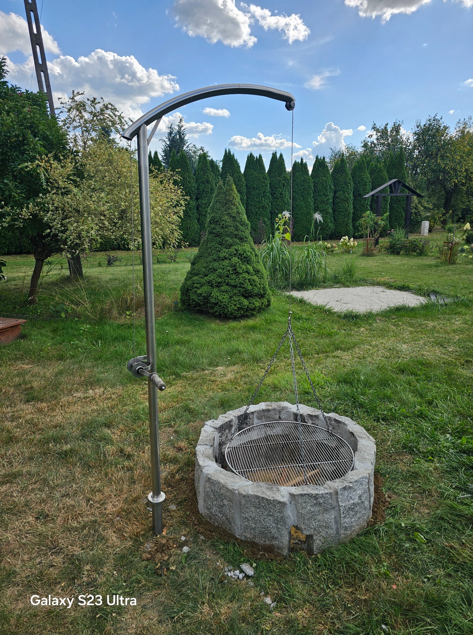 Metalowy grill wiszący nad kamiennym paleniskiem, z mechanizmem regulacji wysokości na tle zadbanego ogrodu i błękitnego nieba z chmurami.