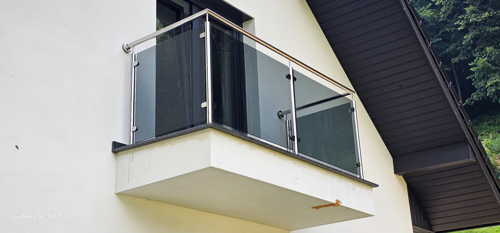 Nowoczesna balustrada ze szkła i stali nierdzewnej na balkonie z kamiennym wykończeniem, widoczna z dołu pod kątem. Tło stanowi elewacja budynku i fragment dachu.