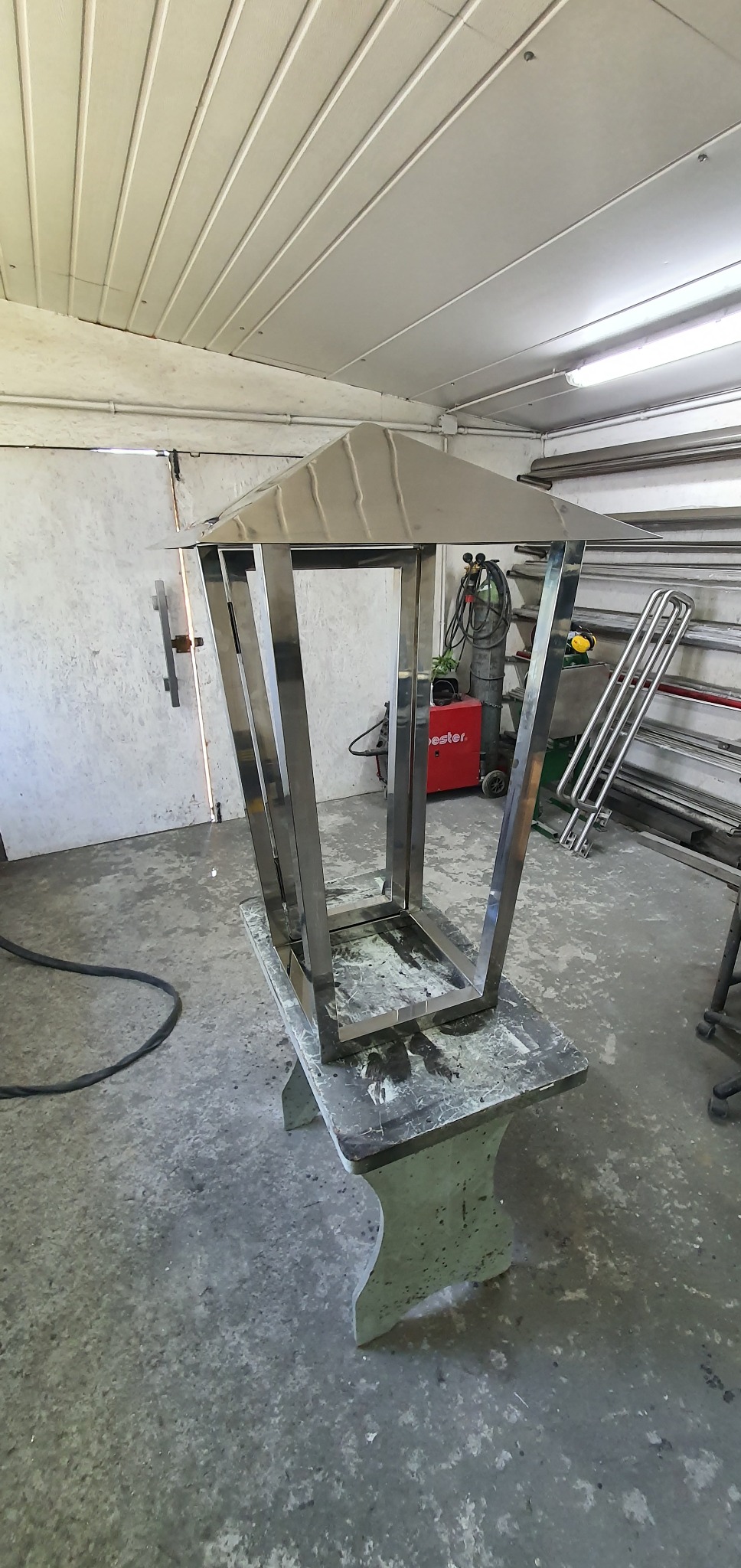 Metalowa konstrukcja przypominająca lampion, wykonana ze stali nierdzewnej, w warsztacie ślusarskim, w trakcie tworzenia. Widoczny spawacz i inne elementy warsztatu.