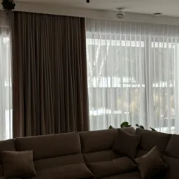 Eleganckie, brązowe zasłony i delikatne, białe firany w przestronnym salonie z dużym narożnikiem, tworzące przytulną atmosferę wnętrza.