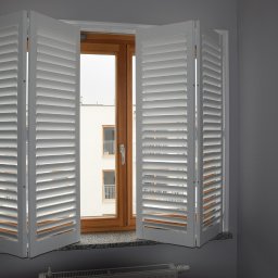 LaurAles Michał Marciniak - Białe, składane okiennice wewnętrzne z regulowanymi lamelami, zamontowane w oknie z drewnianą ramą. Widok na budynek z balkonami przez otwarte okno.