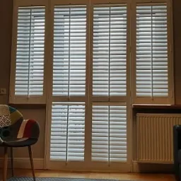 Białe, drewniane okiennice wewnętrzne z regulowanymi lamelami, zamontowane w oknie, widoczne z wnętrza pokoju. Na podłodze szary dywan i krzesło z kolorowym patchworkiem.