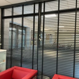 LaurAles Michał Marciniak - Eleganckie, grafitowe żaluzje aluminiowe w biurze, widoczne z perspektywy niskiego kąta, podkreślone czerwonymi fotelami i szklanym stolikiem.