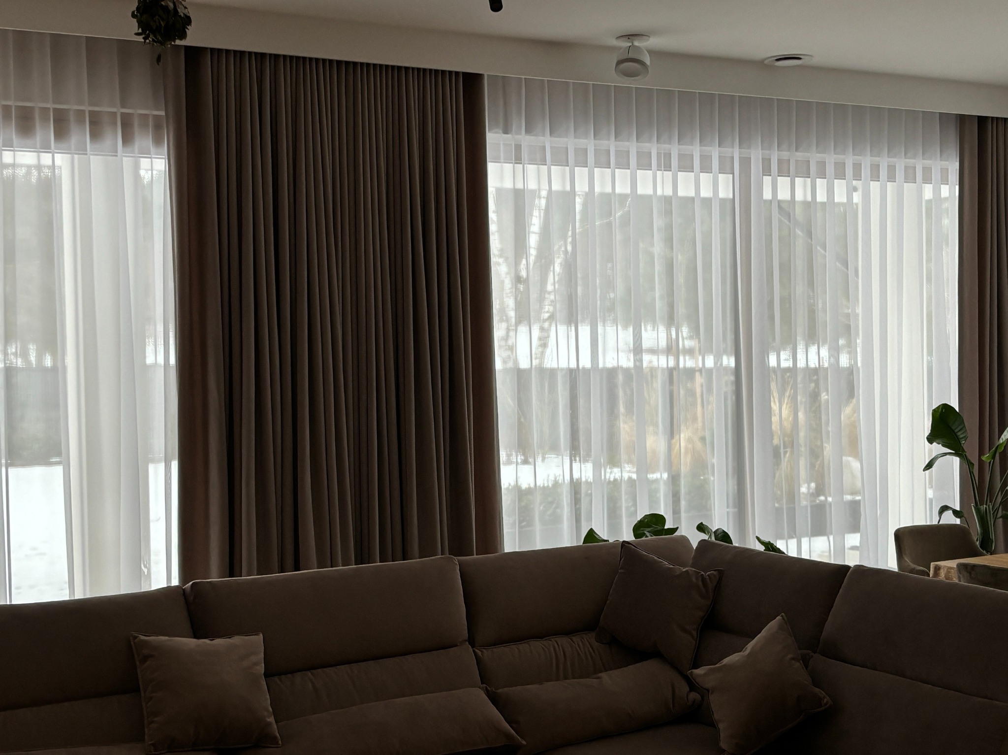 Eleganckie, brązowe zasłony i delikatne, białe firany w przestronnym salonie z dużym narożnikiem, tworzące przytulną atmosferę wnętrza.