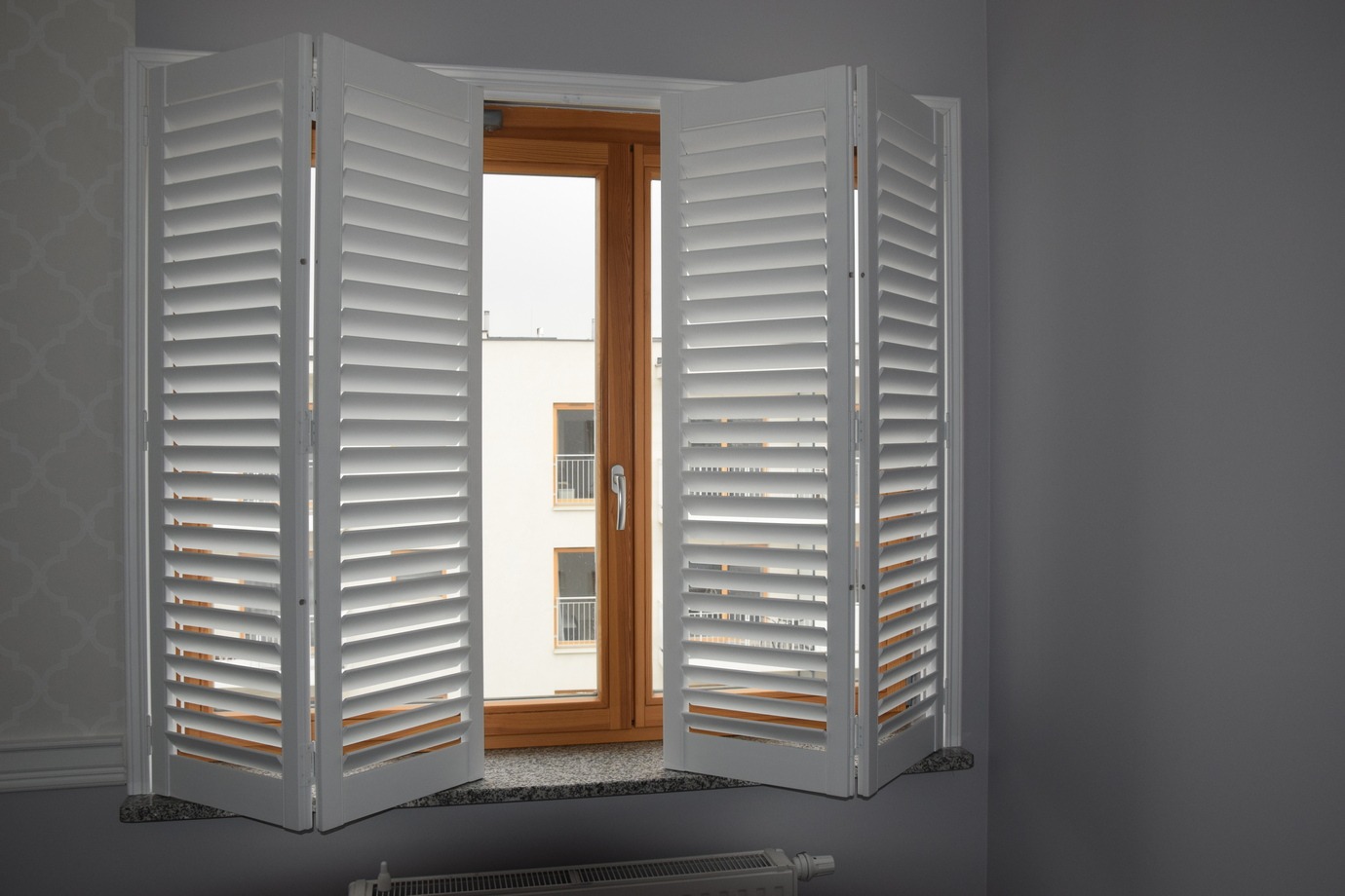 Białe, składane okiennice wewnętrzne z regulowanymi lamelami, zamontowane w oknie z drewnianą ramą. Widok na budynek z balkonami przez otwarte okno.