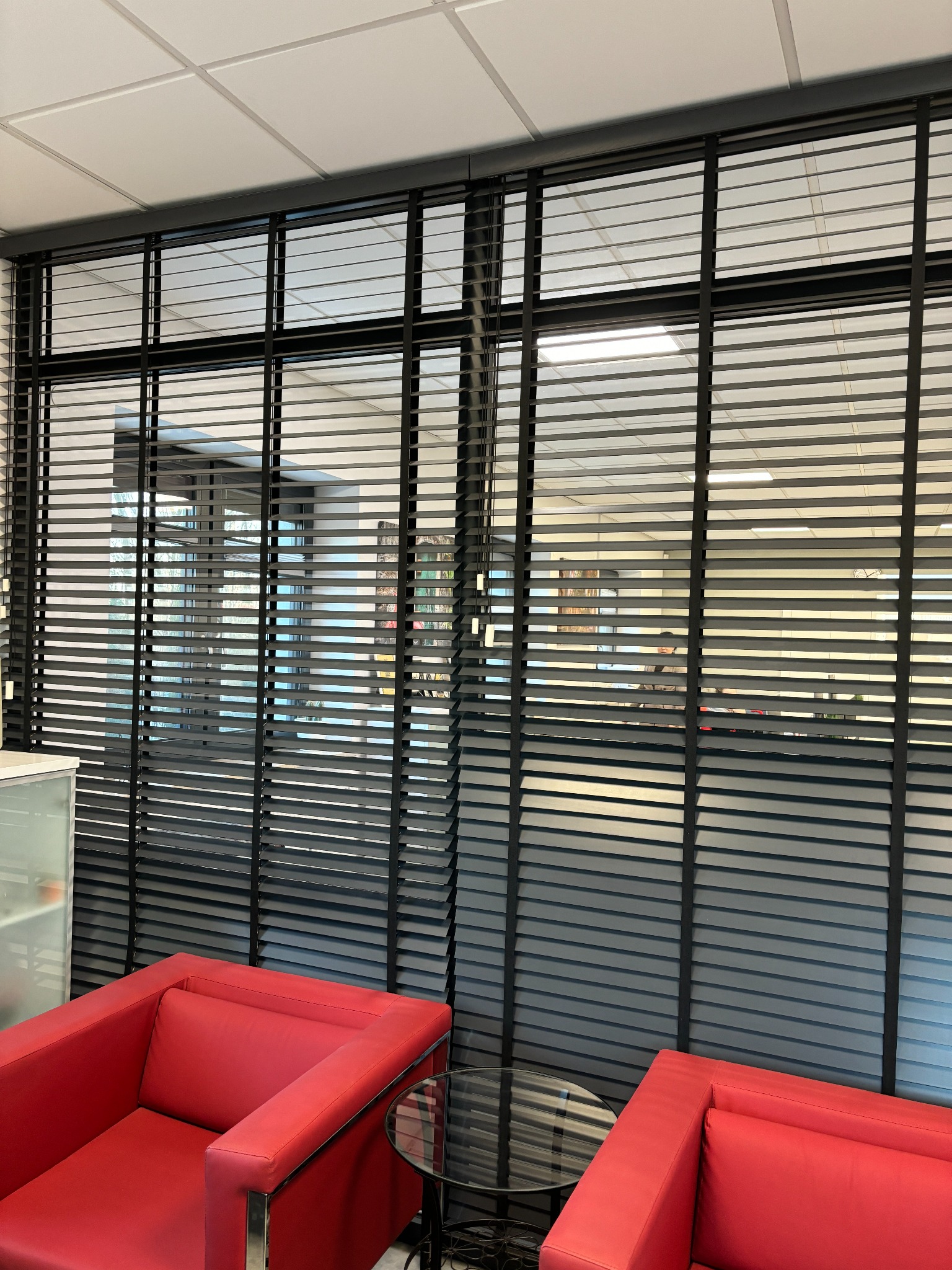 Eleganckie, grafitowe żaluzje aluminiowe w biurze, widoczne z perspektywy niskiego kąta, podkreślone czerwonymi fotelami i szklanym stolikiem.