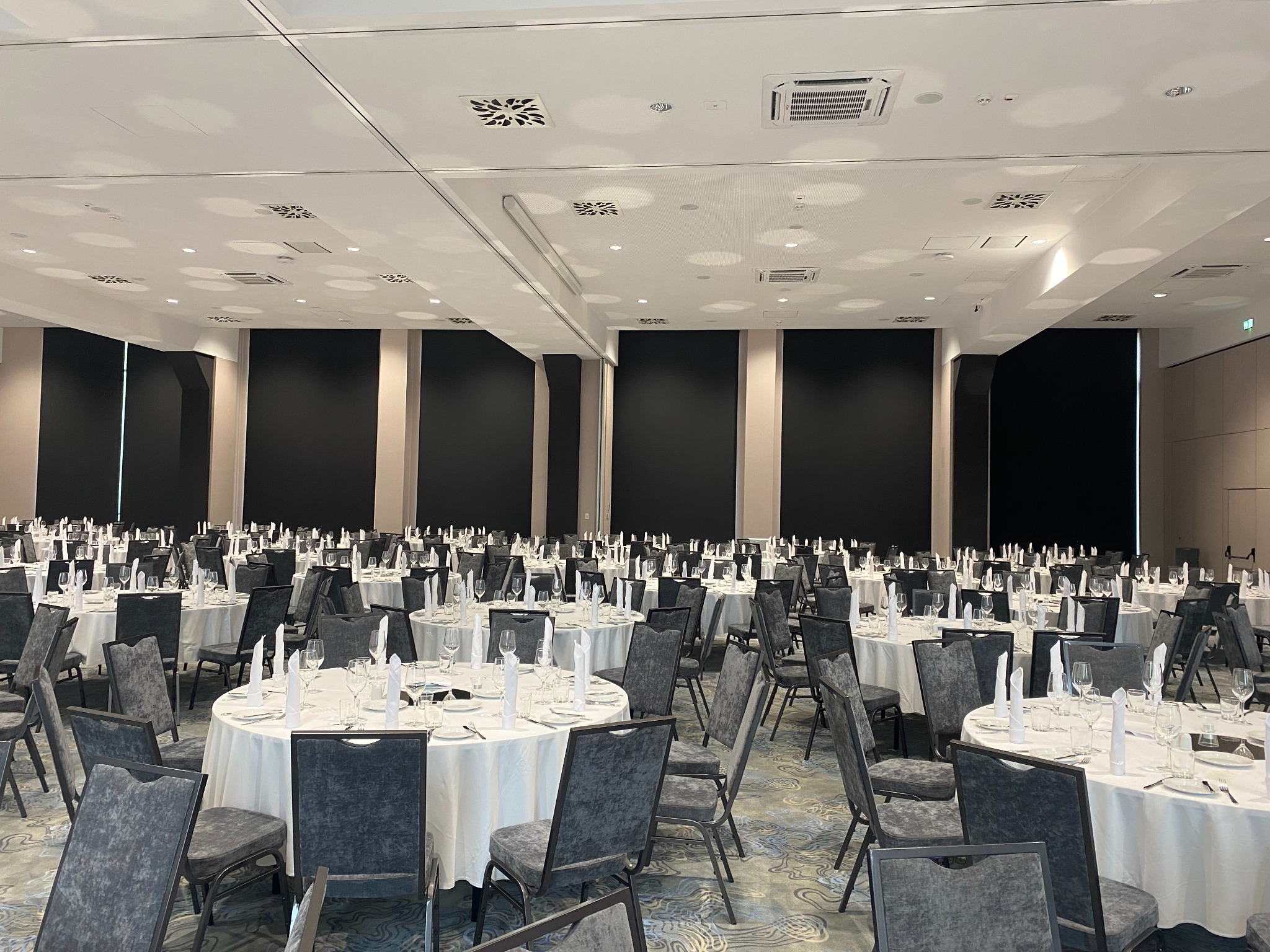 Sala bankietowa z okrągłymi stołami i szarymi krzesłami, przygotowana na uroczystość. Sufit z klimatyzacją i oświetleniem punktowym. Minimalistyczny wystrój.