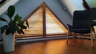 Trójkątne okno strychowe z drewnianymi lamelami, zamykane na środku. Obok fotel i roślina doniczkowa. Parkiet odbija ciepłe światło słoneczne.