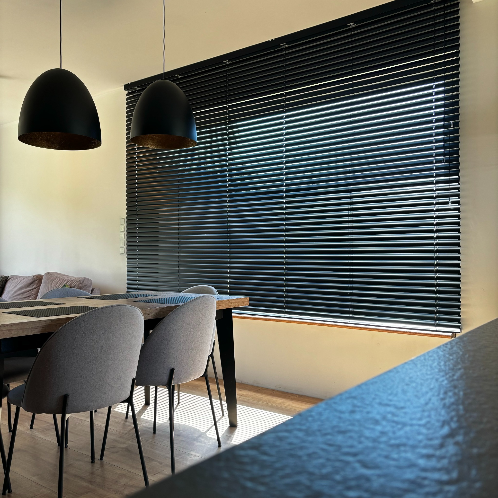 Czarne żaluzje aluminiowe w nowoczesnym wnętrzu jadalni, z widocznymi elementami drewnianego stołu i designerskich lamp wiszących. Promienie słoneczne wpadają przez okno.