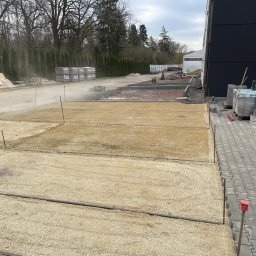 Manus Company Marcin Matusiak - Przygotowany teren pod kostkę brukową: podsypka z piasku, obrzeża betonowe, palety z kostką. Widoczne narzędzia i zraszanie wodą. Częściowo ułożona kostka brukowa.