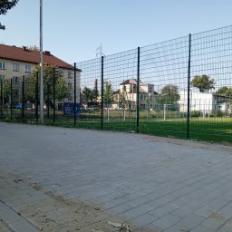 Manus Company Marcin Matusiak - Szara kostka brukowa ułożona na chodniku, z zielonym ogrodzeniem i boiskiem w tle, w słoneczny dzień. Widoczne ślady piasku na kostce.