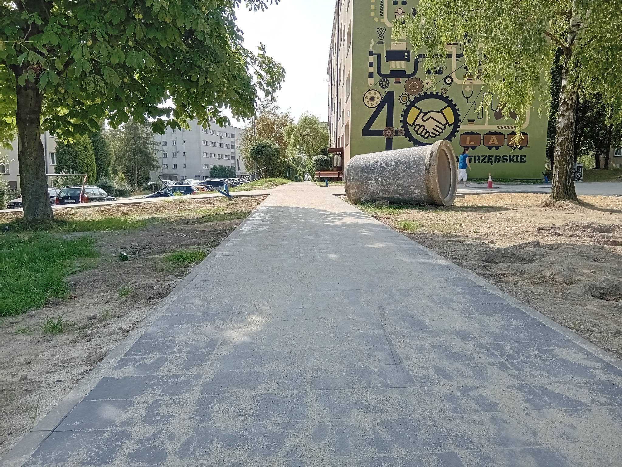 Ułożona ścieżka z kostki brukowej prowadząca do budynku z muralem, z widocznym betonowym elementem dekoracyjnym. Otoczenie zielone, drzewa.