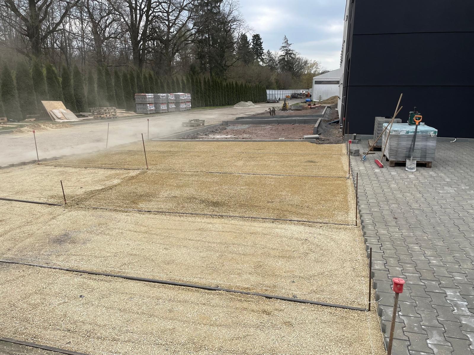 Przygotowany teren pod kostkę brukową: podsypka z piasku, obrzeża betonowe, palety z kostką. Widoczne narzędzia i zraszanie wodą. Częściowo ułożona kostka brukowa.