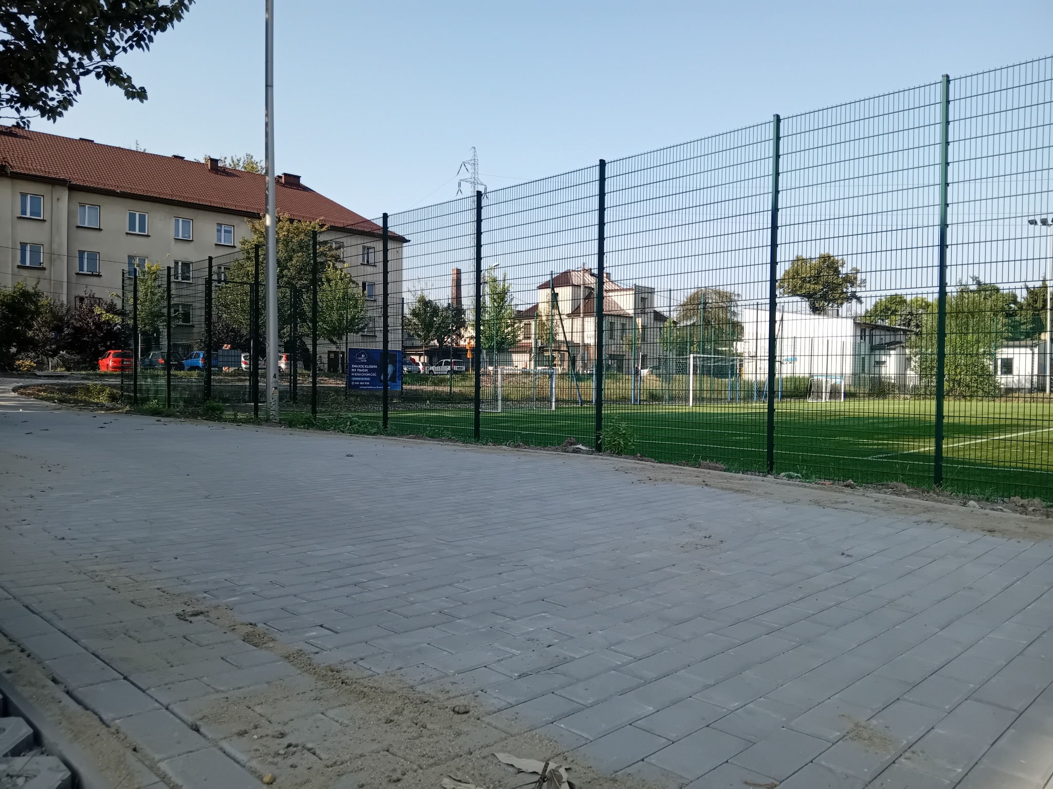 Szara kostka brukowa ułożona na chodniku, z zielonym ogrodzeniem i boiskiem w tle, w słoneczny dzień. Widoczne ślady piasku na kostce.