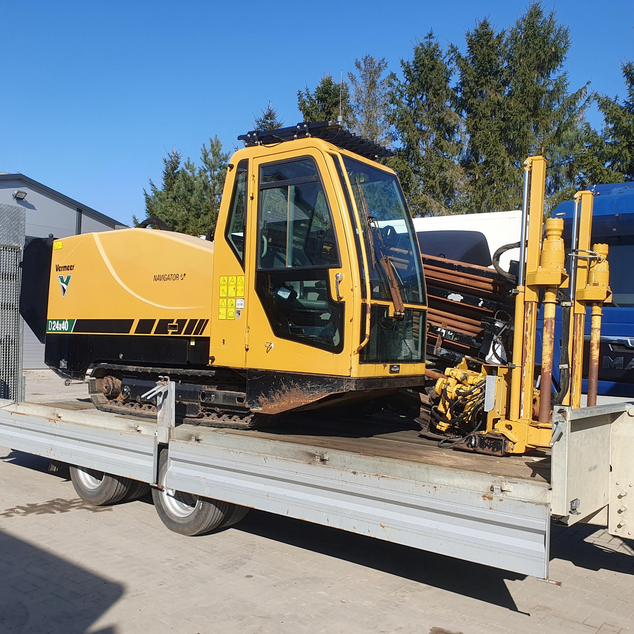Żółta wiertnica horyzontalna Vermeer Navigator D24x40 na lawecie transportowej, widok z boku na tle zielonych drzew i błękitnego nieba.