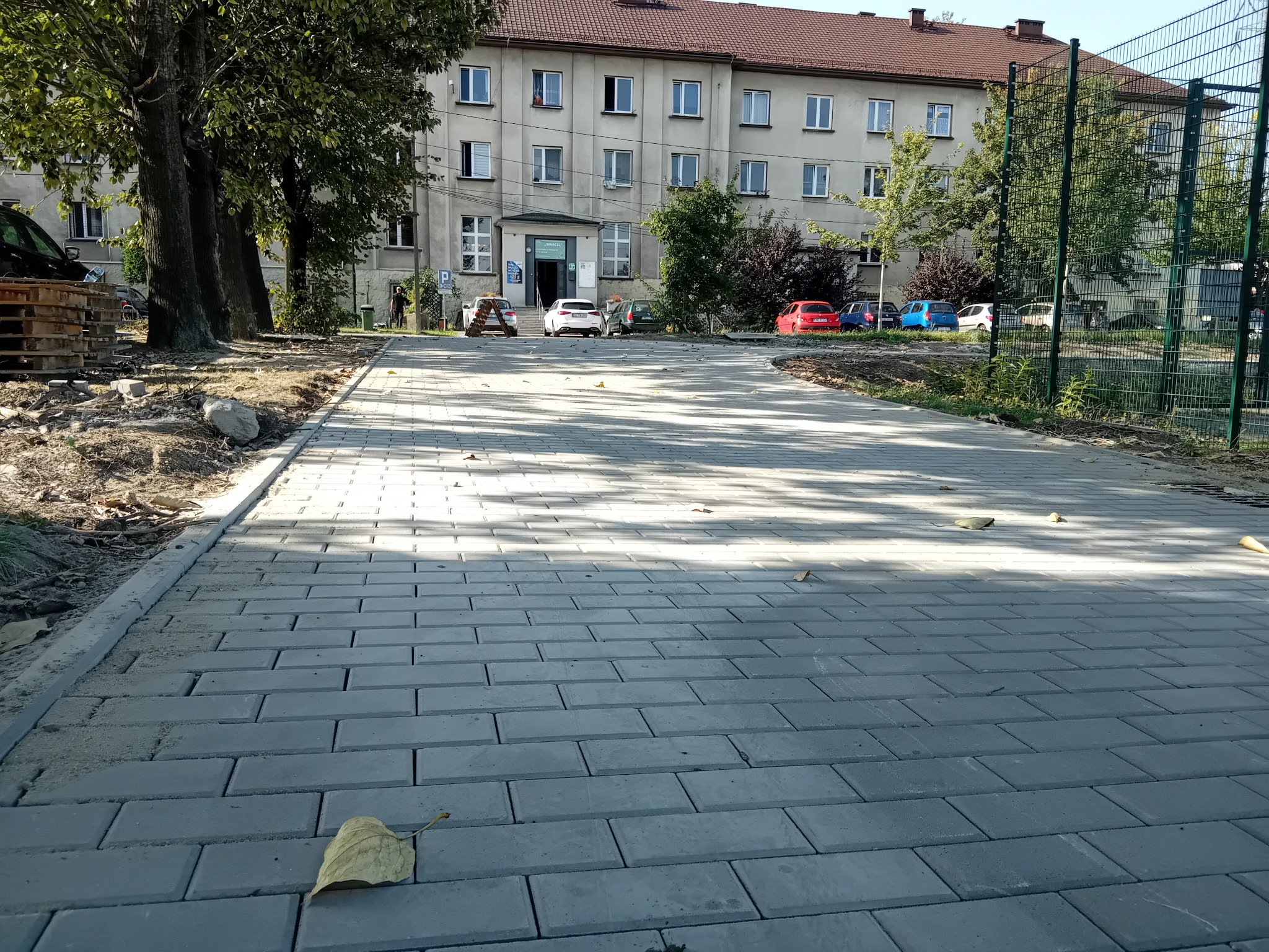 Nowa, szara kostka brukowa ułożona na ścieżce przed budynkiem. Widoczne drzewa, samochody i fragment placu budowy. Na kostce leży pojedynczy, żółty liść.