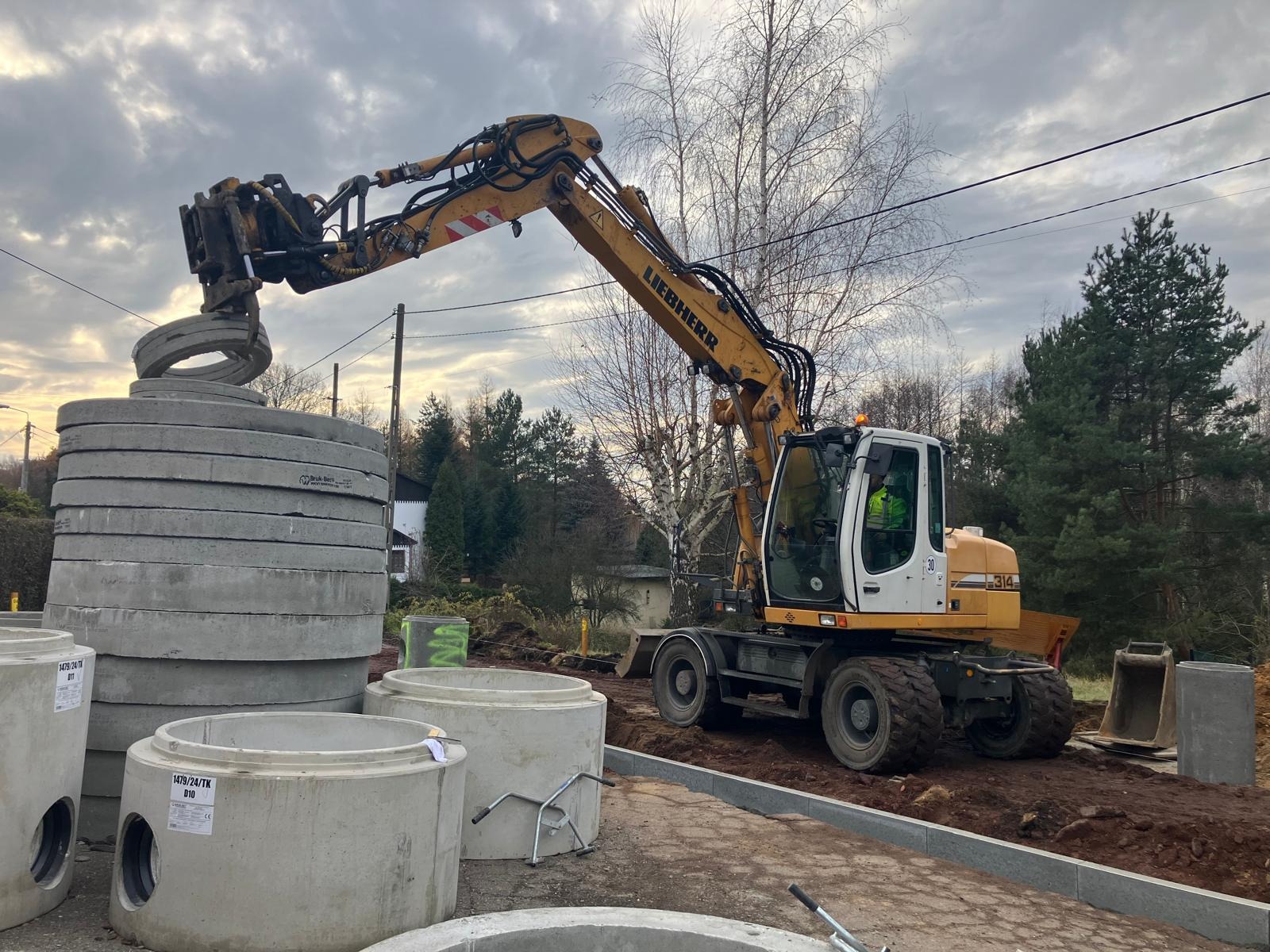 Żółta koparka Liebherr podnosi betonowy krąg na placu budowy. Widoczne są ułożone kręgi i przygotowany teren pod instalację.