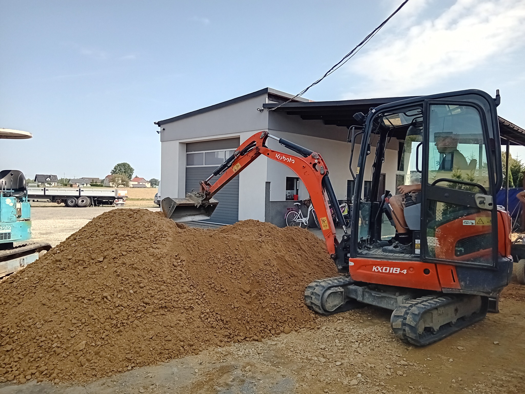 Pomarańczowa mini koparka Kubota KX018-4 z operatorem nabiera ziemię z pryzmy na placu budowy, w tle budynek z bramą garażową i rowery.