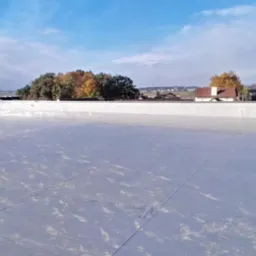 Szary, płaski dach pokryty membraną PVC, z widocznymi zgrzewami, zabezpieczony białym pasem blachy na krawędzi. W tle zabudowa miejska i drzewa.