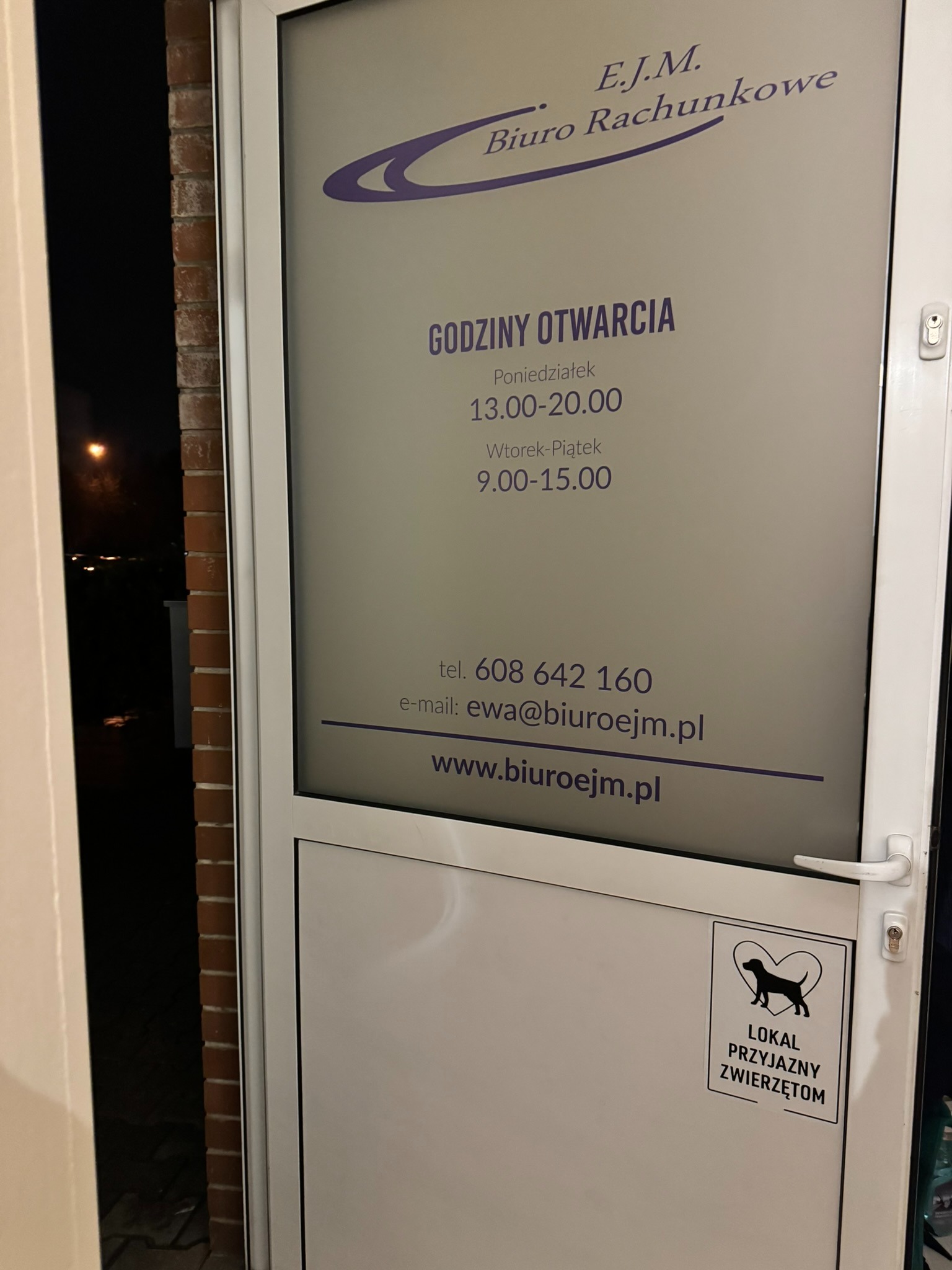 Wejście do biura rachunkowego E.J.M. z godzinami otwarcia na szybie i naklejką 'Lokal przyjazny zwierzętom' na drzwiach. Widoczne dane kontaktowe i logo firmy.