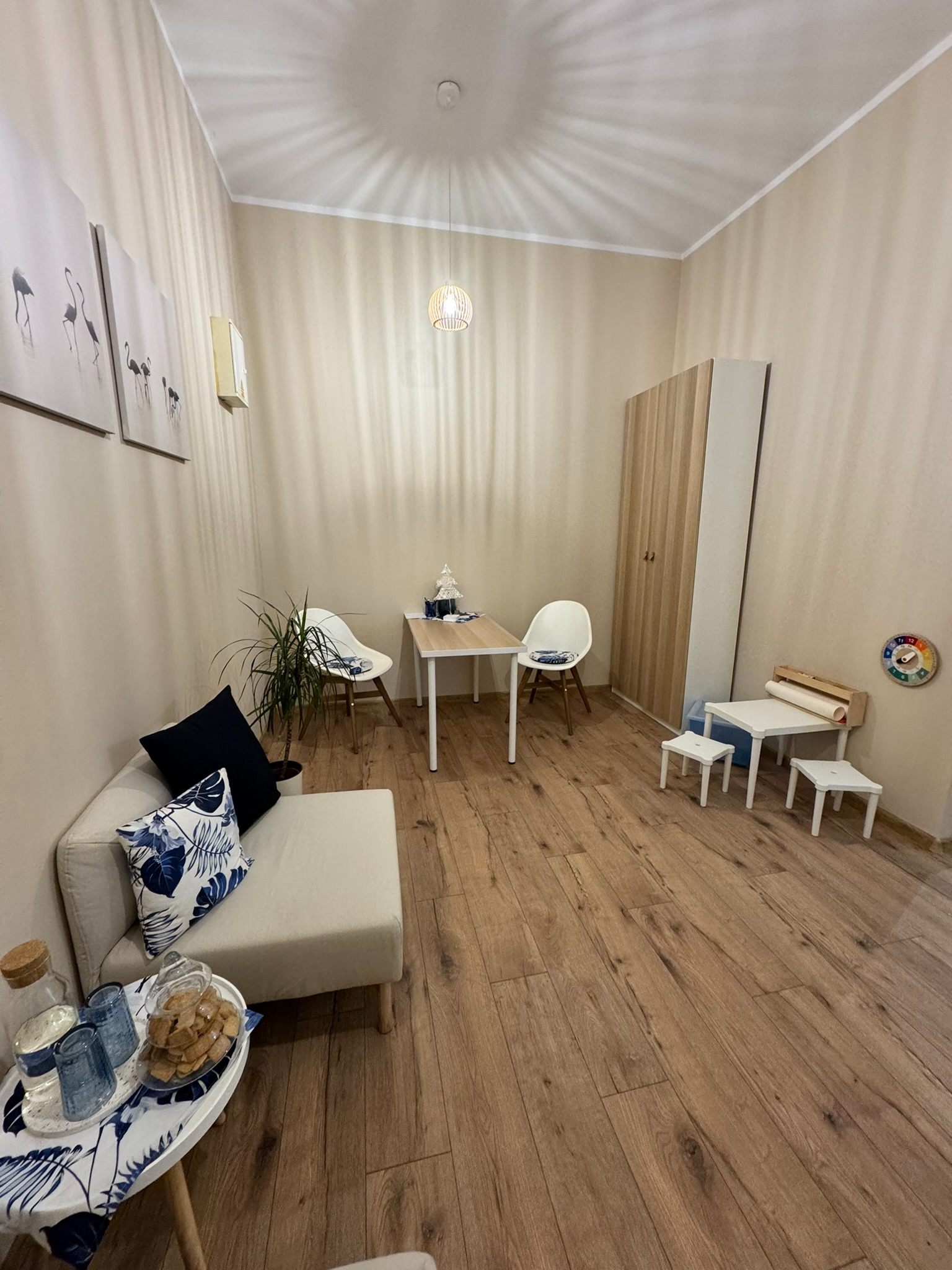 Jasne, przytulne biuro z drewnianą podłogą. W tle biurko, krzesła i szafa. Na pierwszym planie stolik z przekąskami i napojami. Na ścianach obrazy z motywem flamingów.