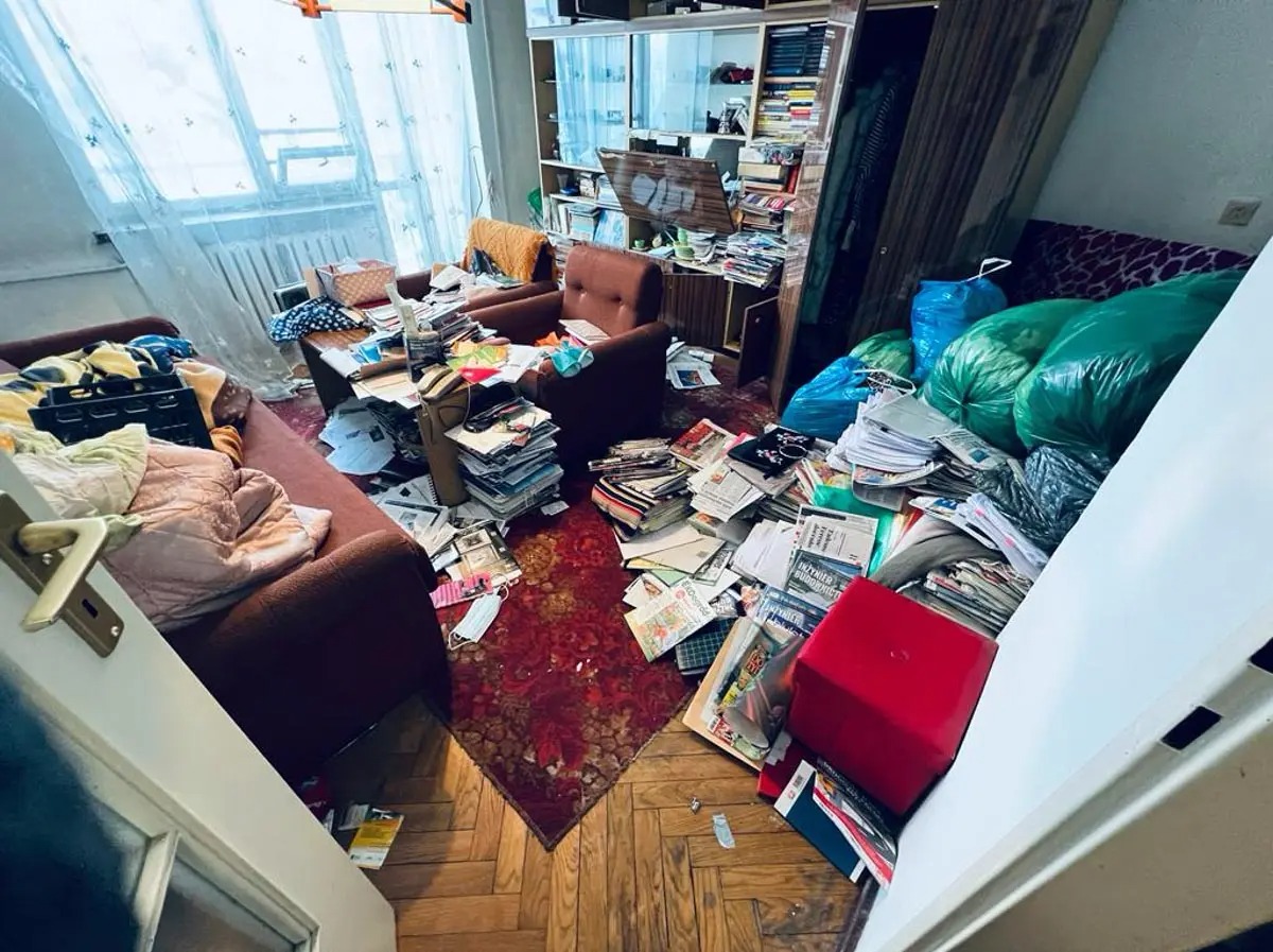 Pokój zagracony stertami gazet, książek i przedmiotów, z widocznymi workami na śmieci, sugerujący potrzebę posprzątania i opróżnienia mieszkania.