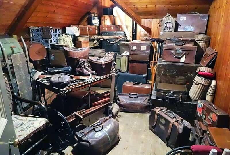 Strych wypełniony starymi walizkami, kuframi i przedmiotami, tworzącymi gęsty, chaotyczny, ale intrygujący widok.