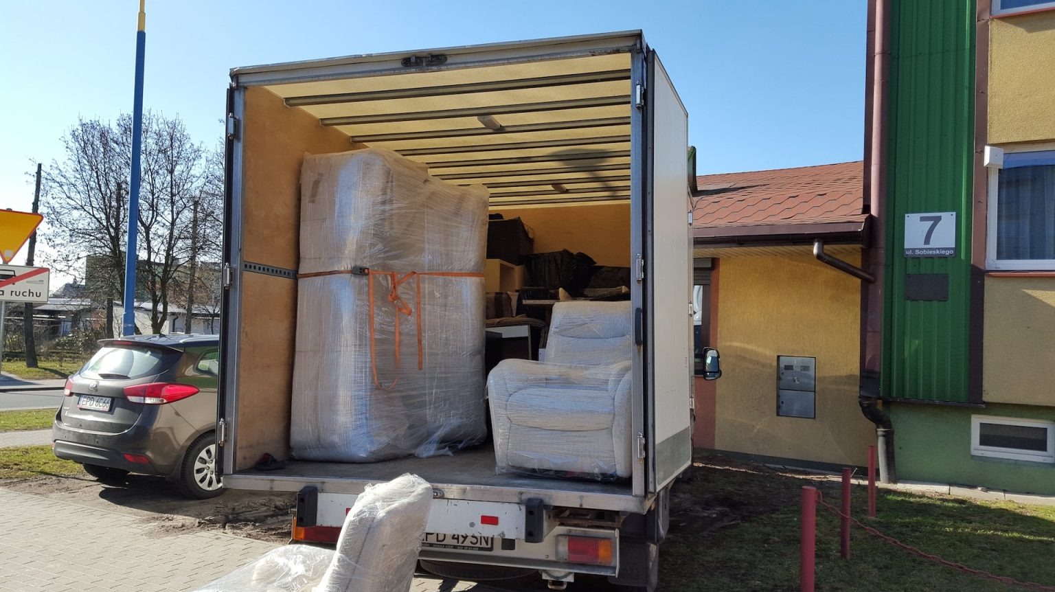 Mebel w folii ochronnej wewnątrz otwartej furgonetki na tle budynku przy ul. Sobieskiego 7. Transport mebli podczas przeprowadzki lub opróżniania.