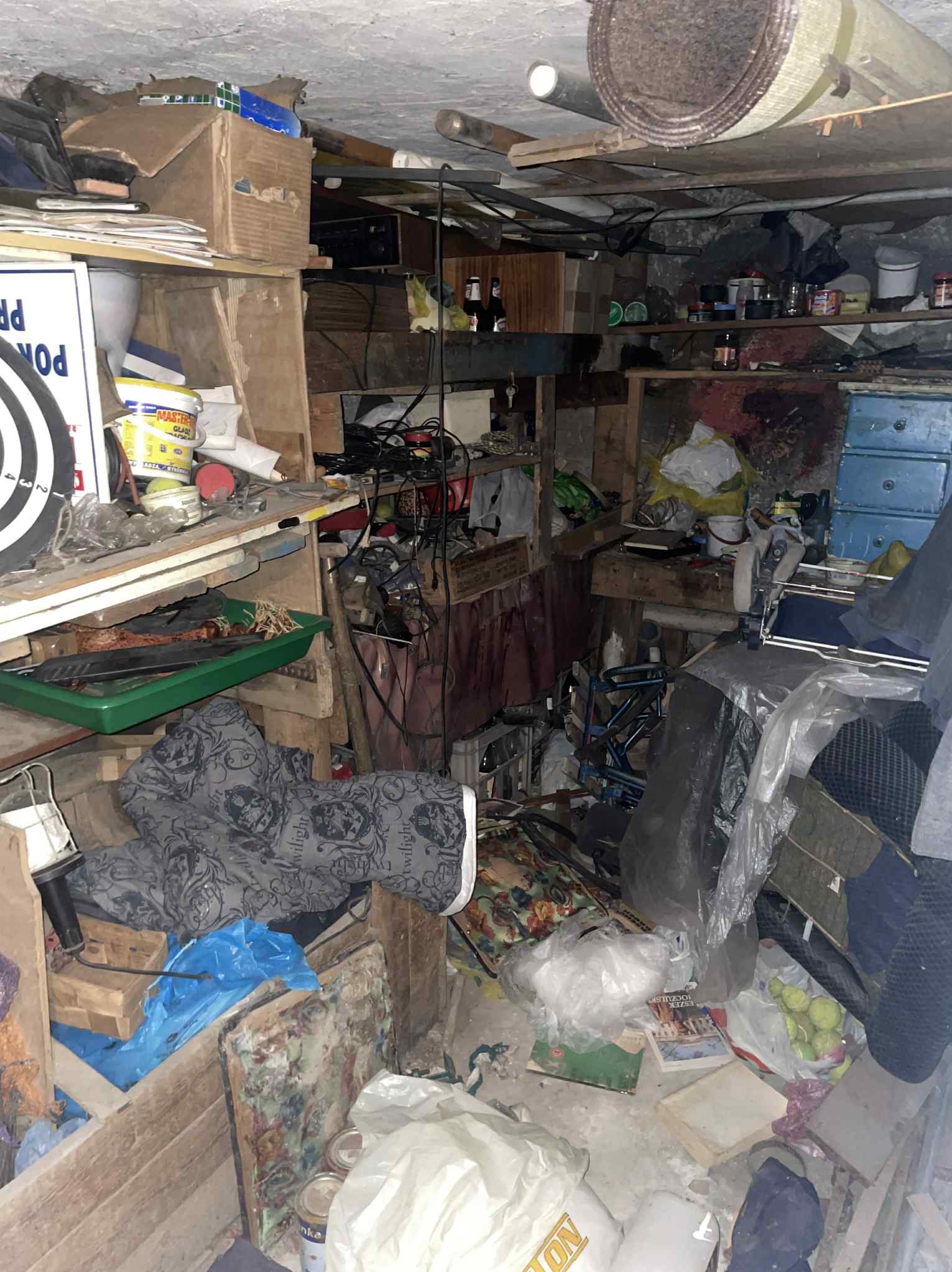 Zastawiony stary pokój z meblami, kartonami, sprzętami i różnymi przedmiotami. Widoczne regały, krzesło inwalidzkie, pudła, butelki i inne przedmioty.