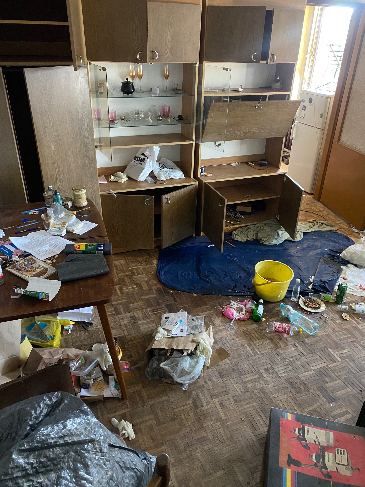 Zaniedbane wnętrze pokoju z otwartymi szafkami, porozrzucanymi przedmiotami, śmieciami i brudną podłogą. Widoczny stół z nieporządkiem i żółte wiadro na podłodze.