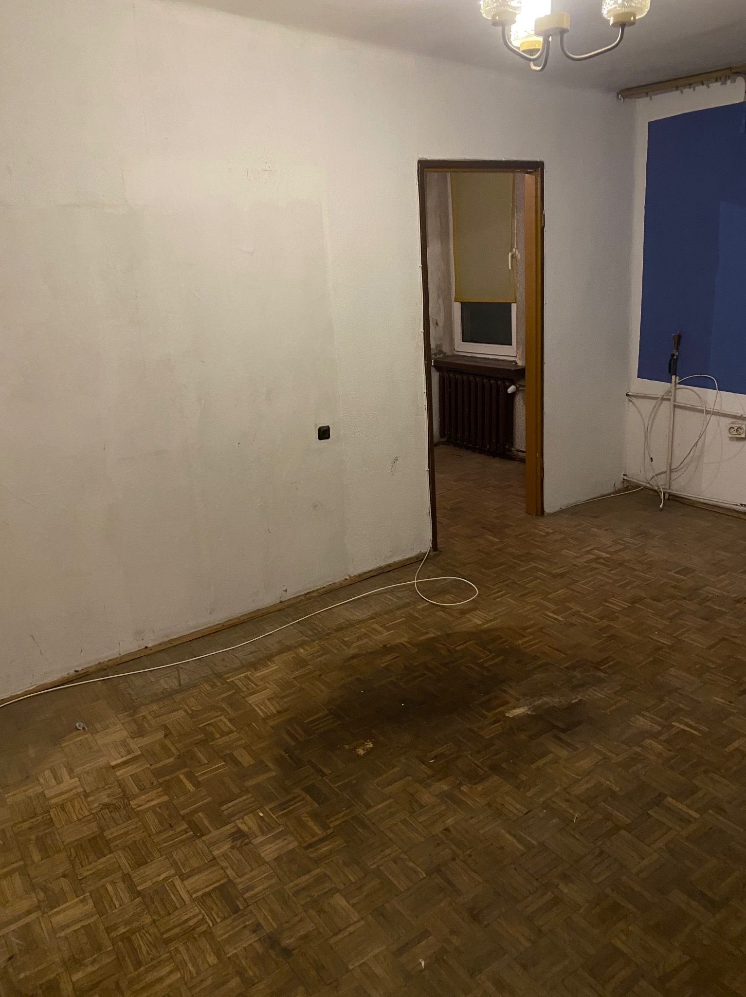 Pusty pokój z parkietem w jodełkę, widoczne ślady użytkowania. Białe ściany, okno z roletą i grzejnik w tle. Kabel na podłodze, lampa sufitowa.