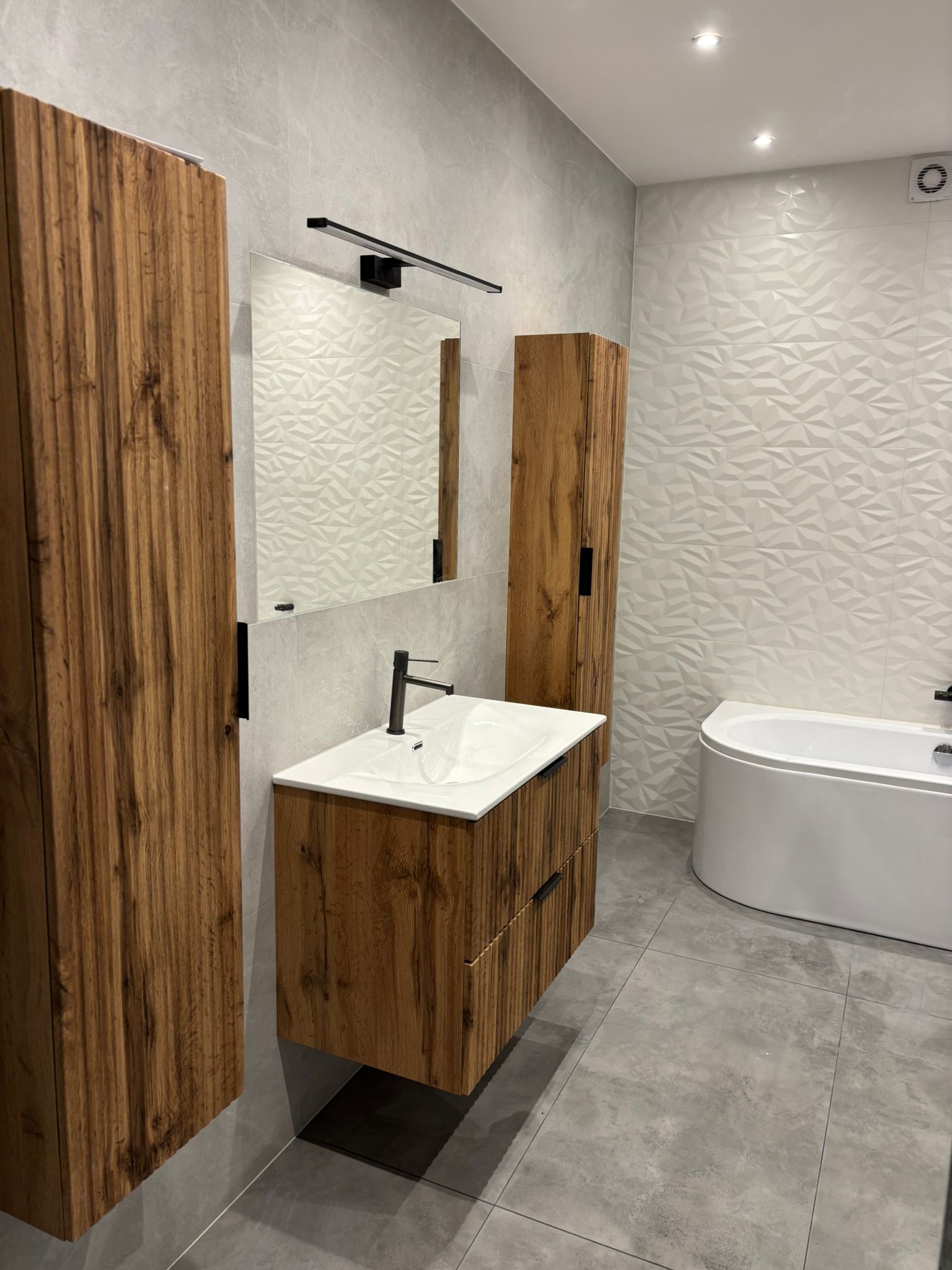 Nowoczesna łazienka z drewnianymi szafkami, umywalką nablatową i geometrycznymi płytkami. Minimalistyczne oświetlenie i szara kolorystyka tworzą elegancki wystrój.