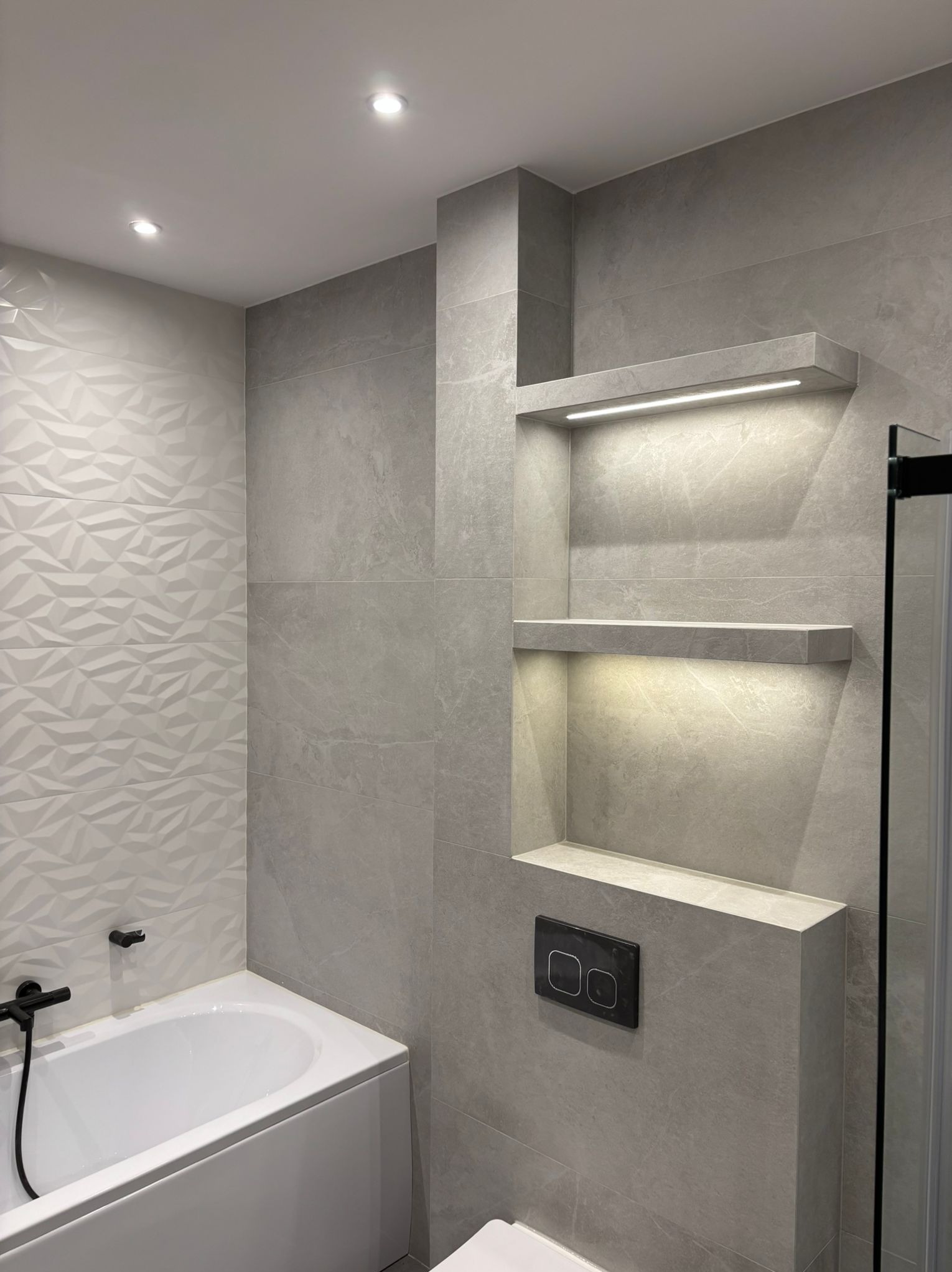 Nowoczesna łazienka z szarymi płytkami, podświetlanymi półkami wnękowymi i białą wanną z czarną armaturą. Geometryczne płytki 3D na ścianie dodają unikalnego charakteru.