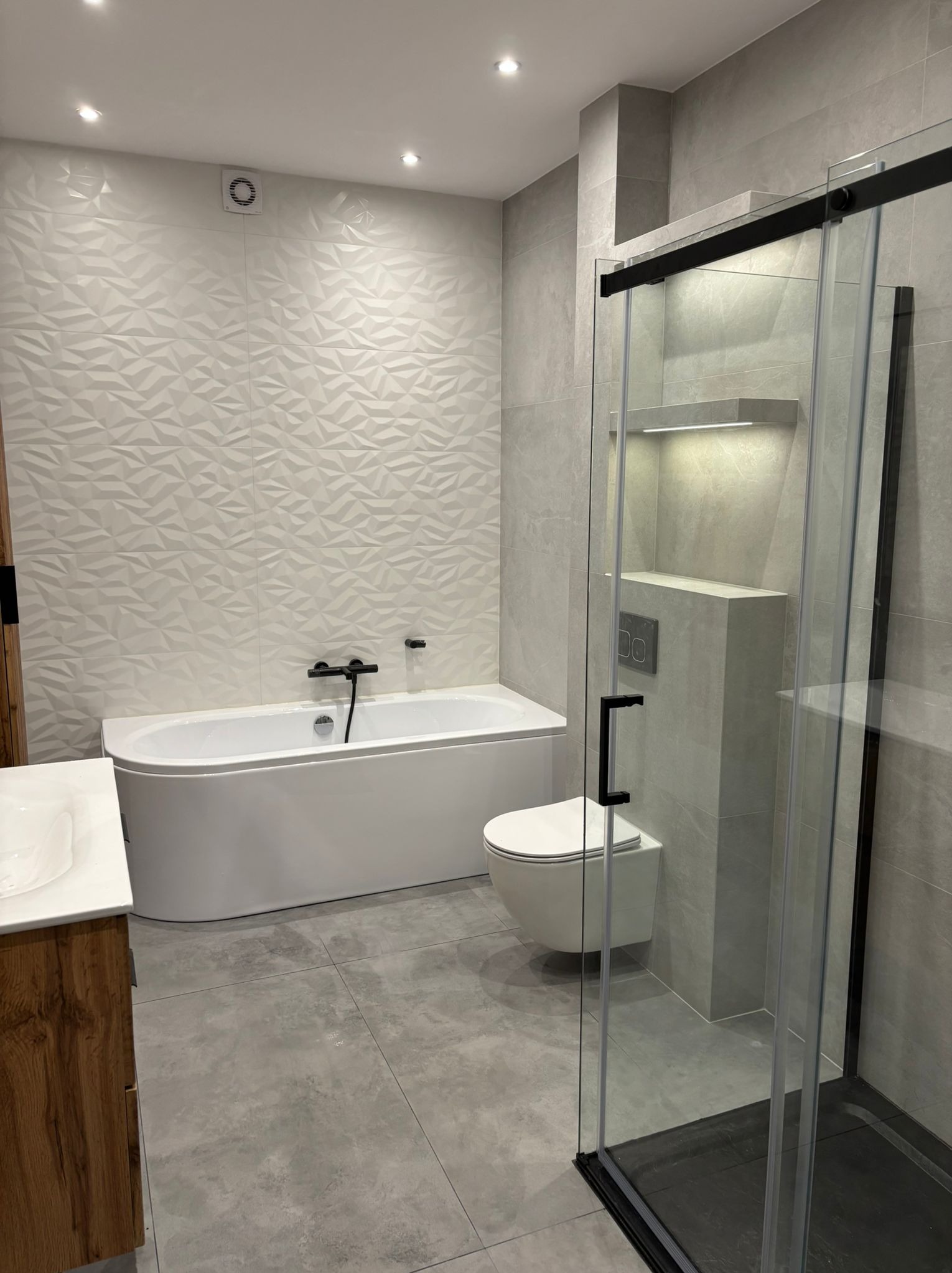 Nowoczesna łazienka z wanną, umywalką w drewnianej szafce, toaletą i kabiną prysznicową z czarnymi detalami. Ściany wyłożone płytkami w odcieniach szarości.