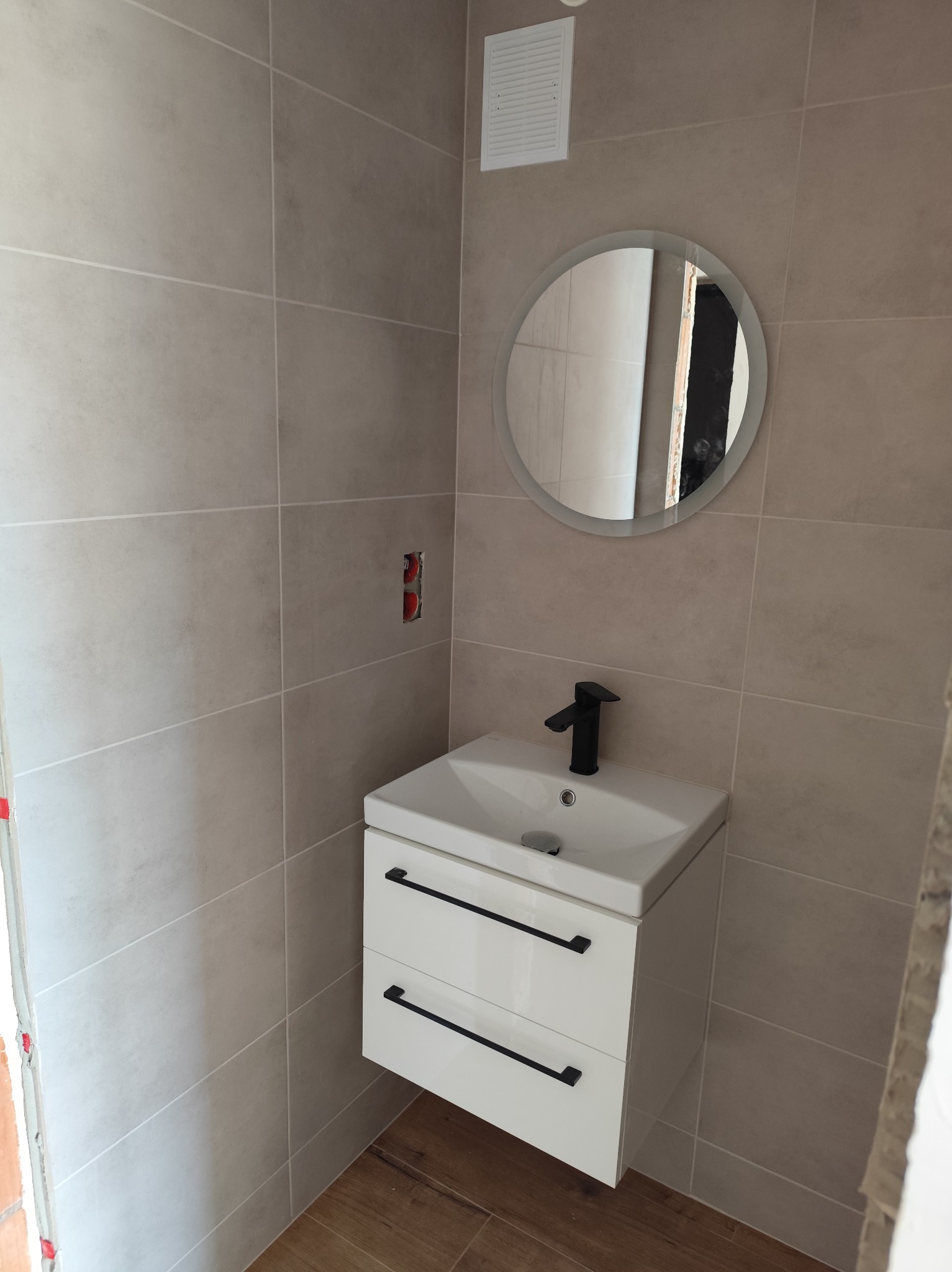 Jasna łazienka z umywalką na szafce z czarnymi uchwytami, okrągłym lustrem i beżowymi płytkami. Widoczna wentylacja i fragment instalacji elektrycznej.