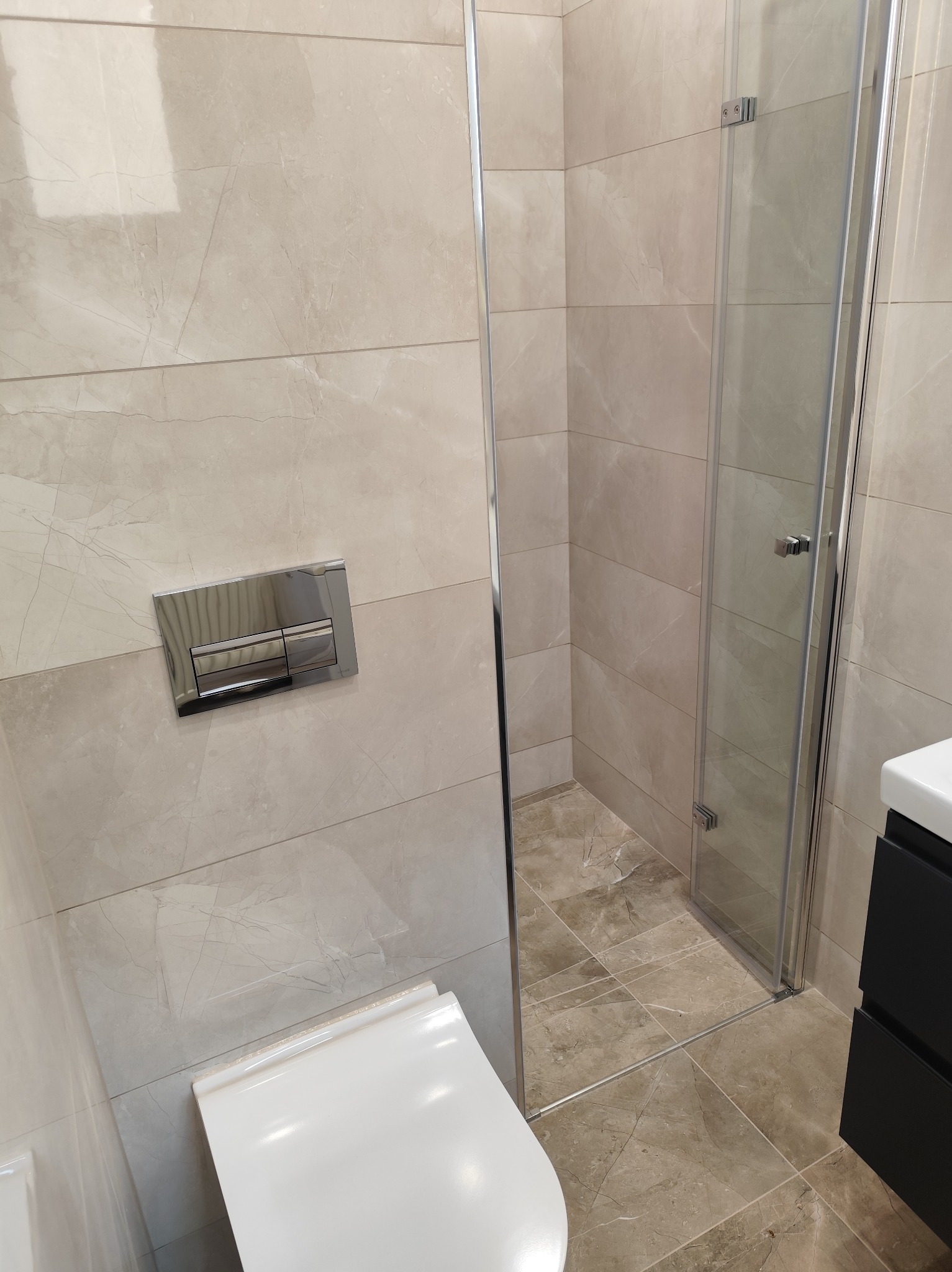 Beżowa łazienka z kabiną prysznicową i podwieszaną toaletą. Płytki imitujące marmur na ścianach i podłodze. Chromowana armatura i czarna szafka pod umywalką.