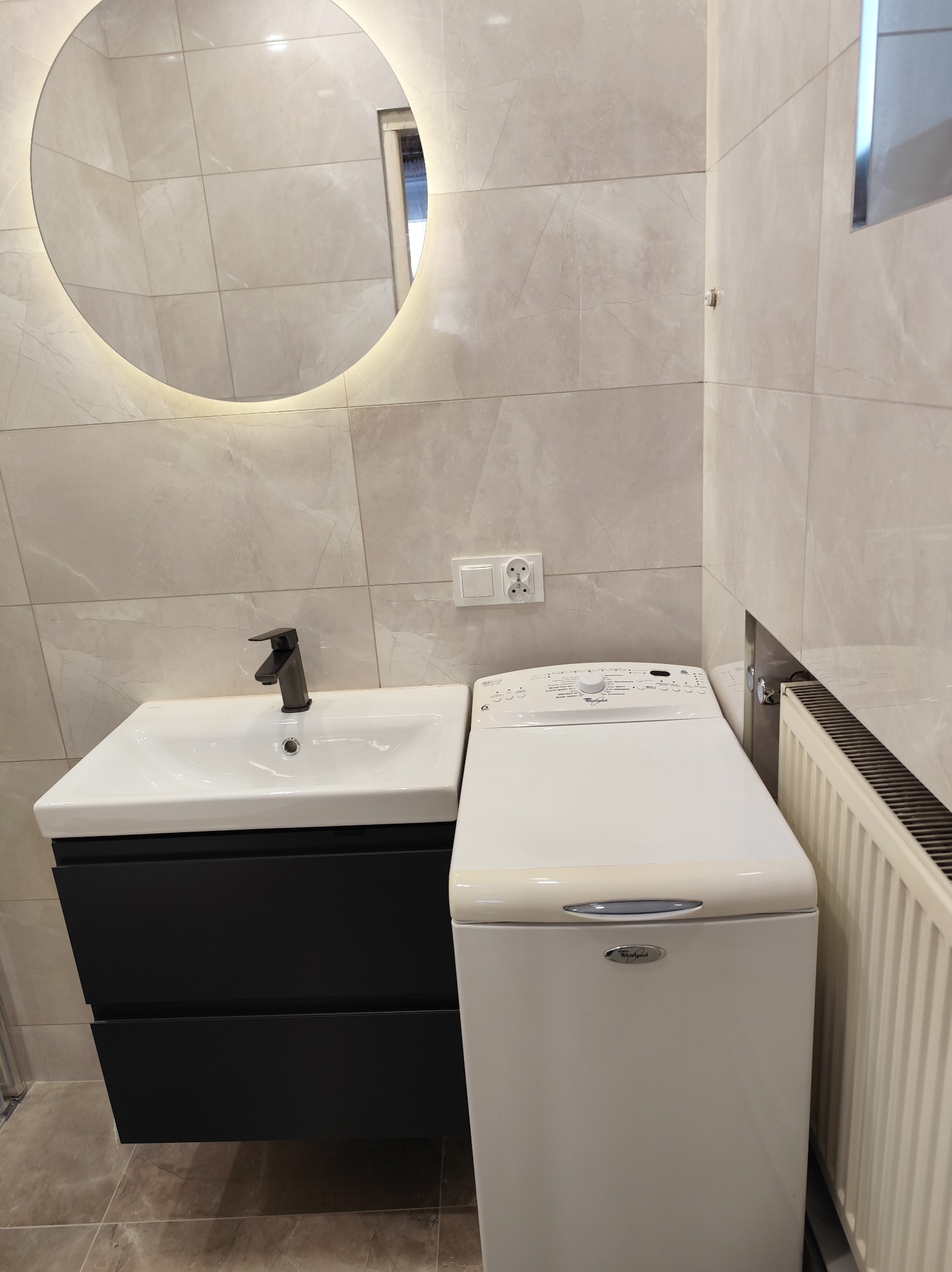 Jasna łazienka z okrągłym lustrem z podświetleniem LED, umywalką z czarną szafką i pralką. Płytki imitujące marmur na ścianach i podłodze. Nowoczesny design.