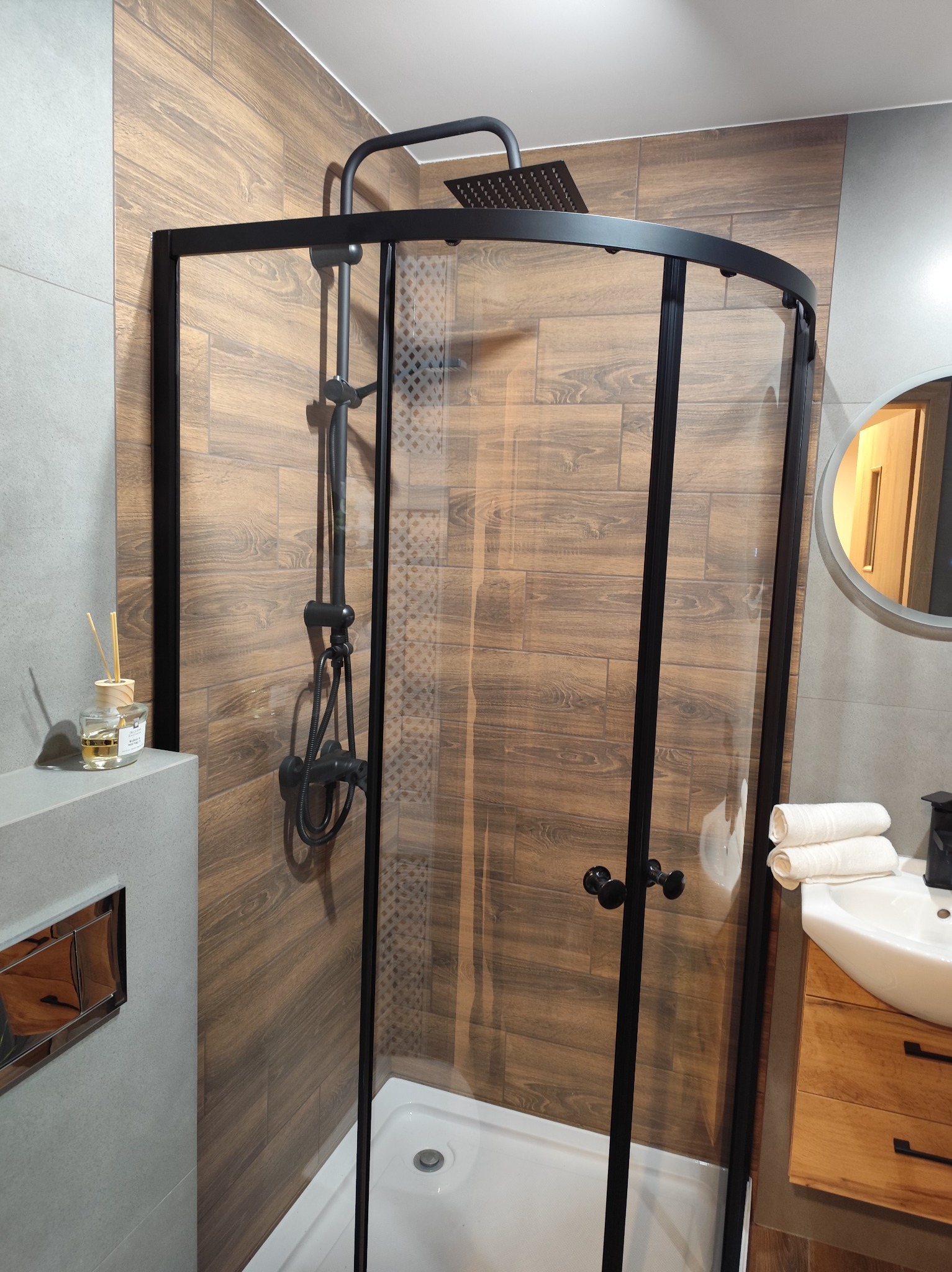 Wykończona łazienka z kabiną prysznicową typu walk-in z czarnymi profilami, drewnopodobne płytki na ścianach, umywalka z szafką z naturalnego drewna.