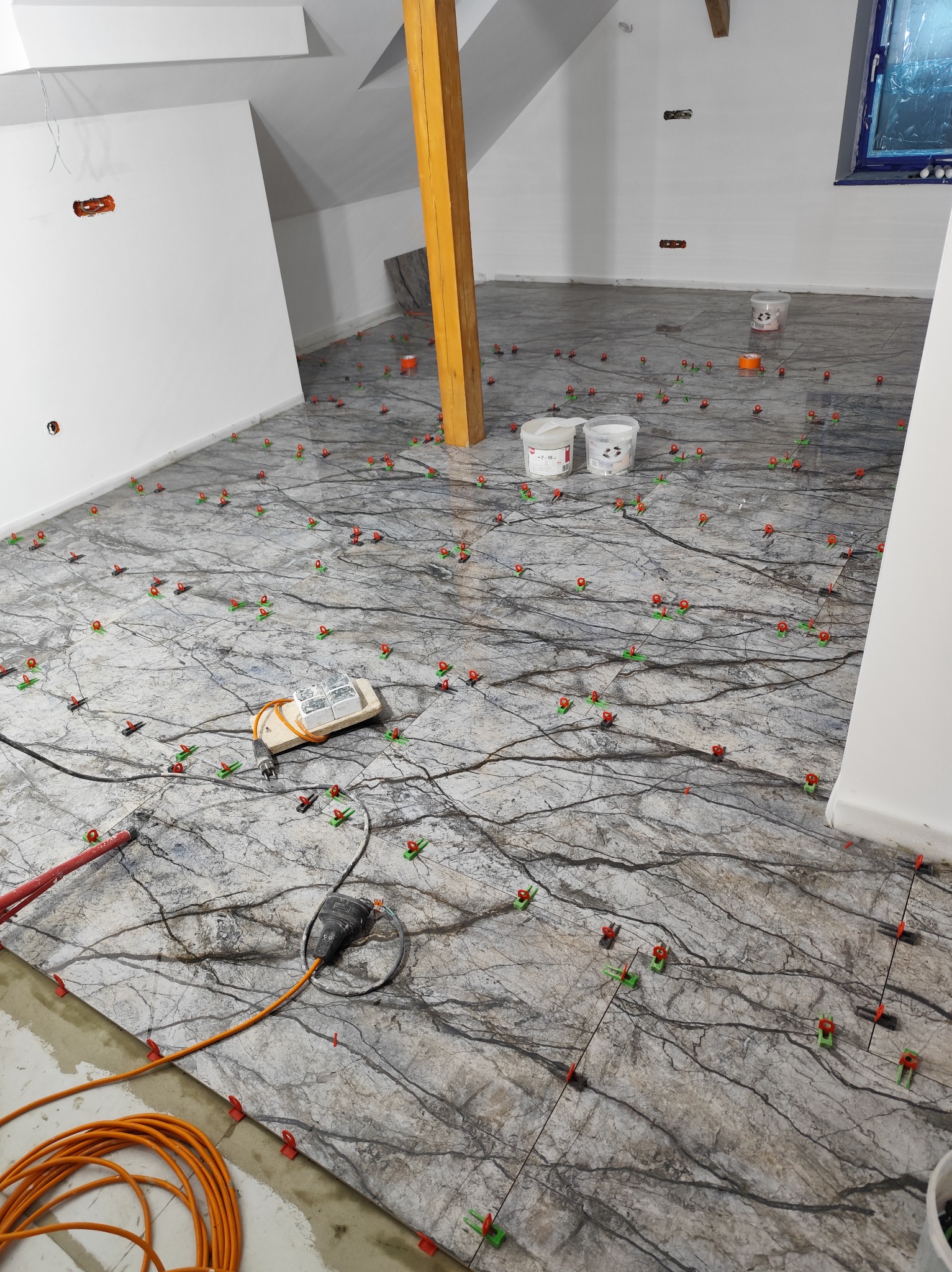 Układanie płytek imitujących marmur na poddaszu. Widoczne poziomice, puszki z klejem i system poziomowania płytek. Naturalne światło z okna.