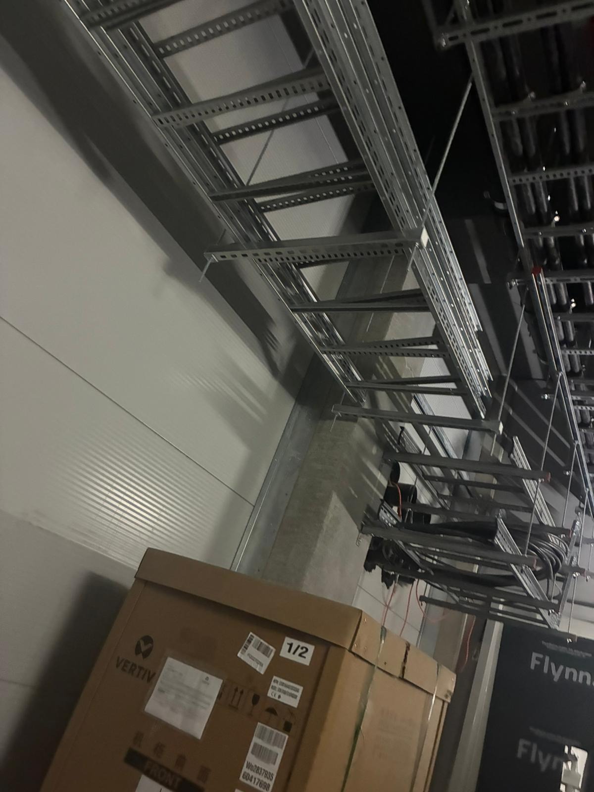 Montaż metalowych korytek kablowych pod sufitem w hali. Widoczne kable i kartony z urządzeniami Vertiv i Flynn. Praca w toku, surowy stan.