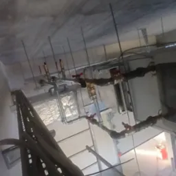 Instalacja rur z zaworami pod sufitem, widoczna drabina i pracownik. Ujęcie z dołu ukazuje skomplikowaną sieć instalacji.