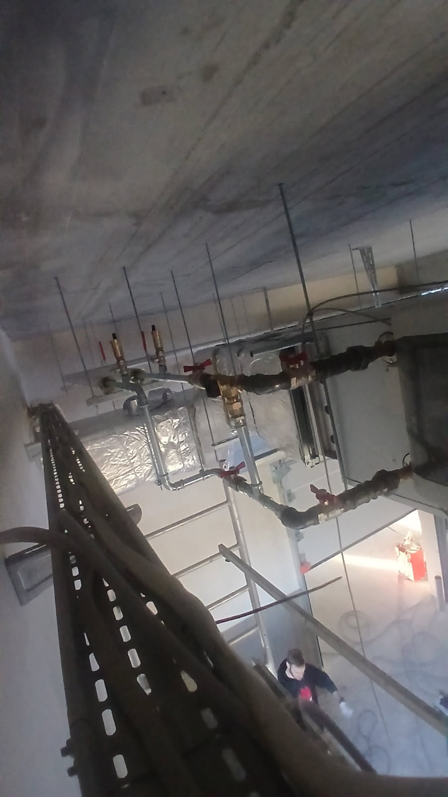 Instalacja rur z zaworami pod sufitem, widoczna drabina i pracownik. Ujęcie z dołu ukazuje skomplikowaną sieć instalacji.
