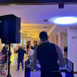 DJ w eleganckiej kamizelce miksuje muzykę na 18-nastce. Parkiet pełen tańczących gości, oświetlenie sceniczne i dekoracja z numerem '18' w tle.