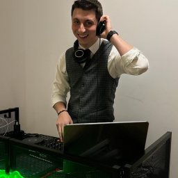 OLSOUND Wiktor Olszewski - Uśmiechnięty DJ w kamizelce, z słuchawkami na szyi, obsługuje konsoletę z neonowym napisem 'Party With SOUND' na tle jasnej ściany.